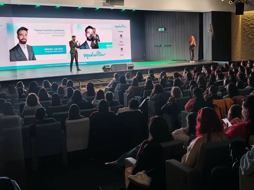 El doctor Miguel Calero, de traje, hablando de pie sobre el escenario de un auditorio con el patio de butacas lleno.